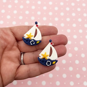 May include: A pair of white and blue sailboat earrings with a yellow star and red accents. The earrings are made of polymer clay.