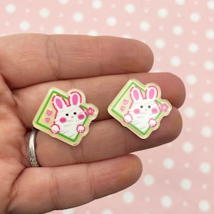Può includere: Due ciondoli in resina a forma di diamante con un coniglietto bianco all'interno. Il coniglietto ha le orecchie rosa e un fiore rosa. Lo sfondo del ciondolo è giallo con linee verdi e cuori rosa.