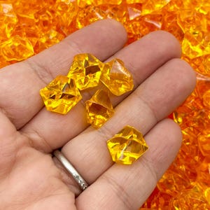May include: A hand holding a handful of small, clear, orange, faceted, plastic gems. The gems are scattered on a surface.