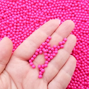 May include: A hand holding a small pile of bright pink round beads. The beads are scattered on a background of more pink beads.
