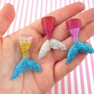 Puede incluir: Tres abalorios de cola de sirena con purpurina en diferentes colores. Las colas est&aacute;n hechas de pl&aacute;stico transparente con purpurina incrustada. Las colas son de color rosa, azul y amarillo.