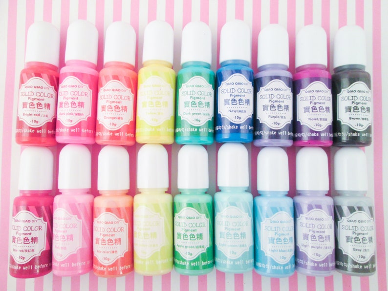 Pu&ograve; includere: Un set di 18 piccoli flaconi di pigmento liquido in vari colori, tra cui rosso, rosa, arancione, giallo, verde, blu, viola, marrone e grigio. Ogni flacone ha un tappo bianco e un'etichetta con il testo "SOLID COLOR Pigment" in inglese e giapponese.