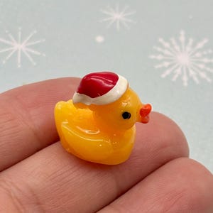May include: A small, yellow rubber ducky wearing a red and white Santa hat. The duck has a bright orange beak and small black eyes. The background is a light blue with white snowflake designs.