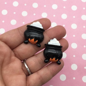 May include: Two black cauldron charms with white steam and orange flames. The charms are made of resin and are approximately 1 inch in size.