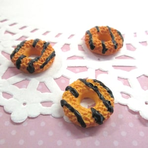 Puede incluir: Tres mini donuts cubiertos de chocolate con glaseado amarillo y rayas de chocolate negro. Los donuts están sobre un encaje blanco sobre un fondo rosa.