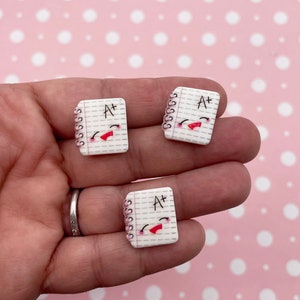 May include: Three white notebook charms with a pink spiral binding and a happy face. Each charm has the text "A+" written on the front.