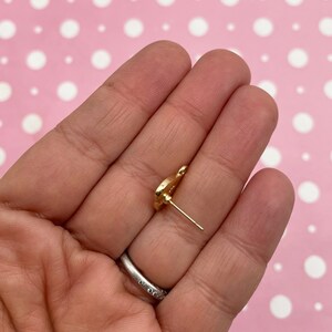 May include: A gold-colored stud earring with a floral design. The earring is held in a hand against a pink background with white polka dots. The earring post is visible.