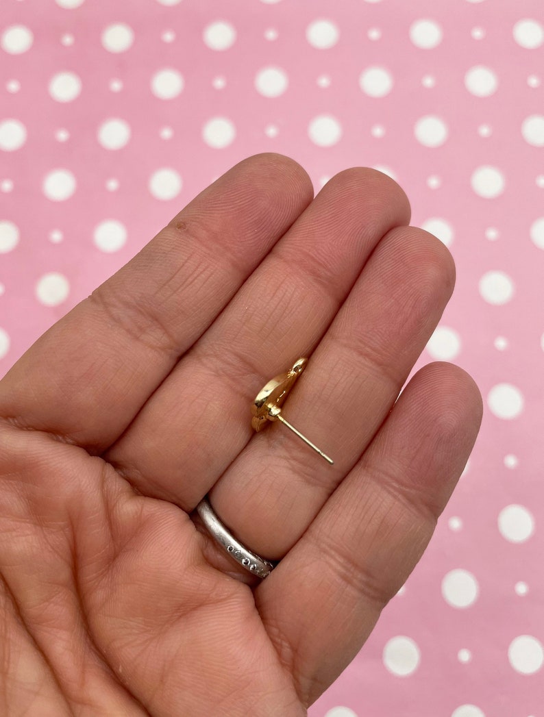 May include: A gold-colored stud earring with a floral design. The earring is held in a hand against a pink background with white polka dots. The earring post is visible.