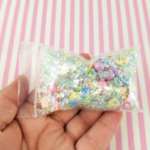 May include: A clear plastic bag filled with iridescent glitter, beads, and small charms in various shapes, including stars, seashells, and hearts. The bag is held in a hand against a pink and white striped background.