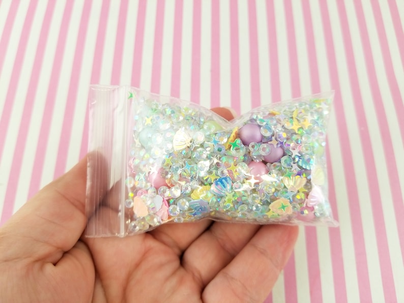 May include: A clear plastic bag filled with iridescent glitter, beads, and small charms in various shapes, including stars, seashells, and hearts. The bag is held in a hand against a pink and white striped background.