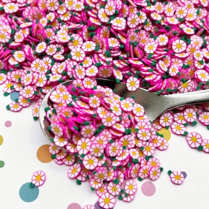 Puede incluir: Un montón de espolvoreados en forma de flor rosa y blanca con hojas verdes. Los espolvoreados están esparcidos sobre una superficie blanca con lunares de colores.