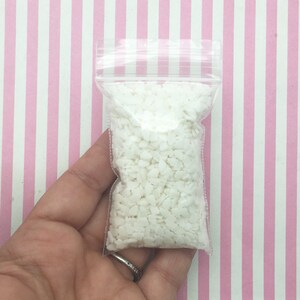 May include: A clear plastic bag filled with small, white, irregularly shaped pieces. The bag is held against a pink and white striped background. The bag is sealed with a zipper closure.