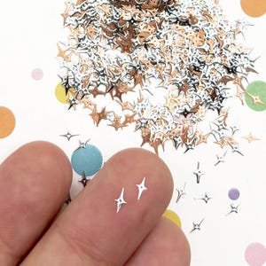 May include: A collection of small, four-pointed star-shaped metal embellishments in silver and rose gold tones. The tiny star charms feature a sharp, geometric design and are scattered across a white surface decorated with colorful polka dots.