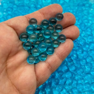 May include: A hand holding a handful of small, translucent blue water beads. The beads are scattered across a surface.