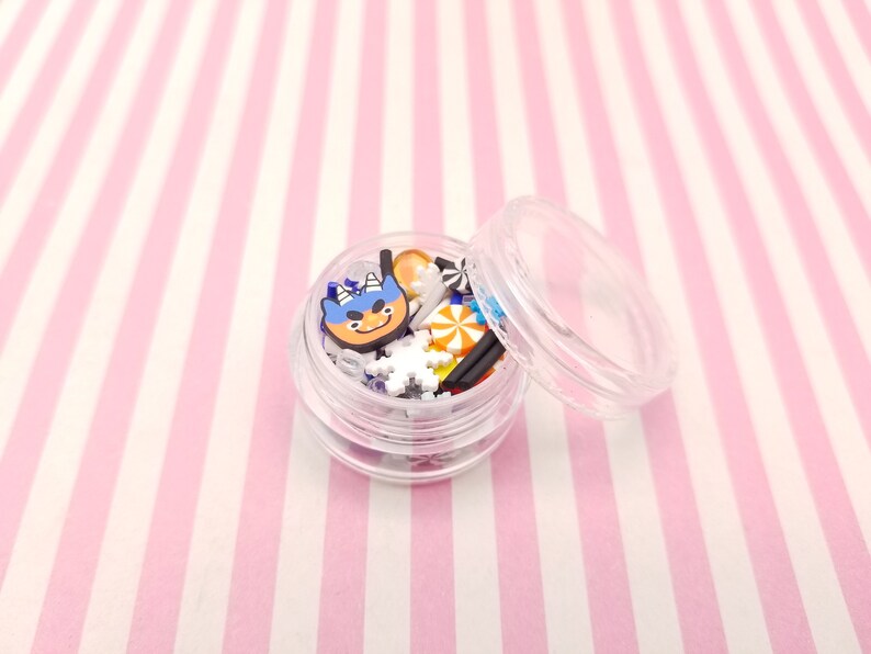 Peut inclure: Un r&eacute;cipient en plastique transparent rempli de charmes en plastique color&eacute;s et blancs, dont un personnage bleu et orange avec un nez rouge, des flocons de neige et des cannes en sucre. Le r&eacute;cipient a un couvercle transparent.