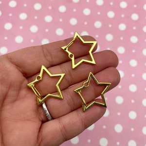 May include: Three gold-colored metal star-shaped charms with a hole at the top. The charms are open and have a smooth, shiny finish.