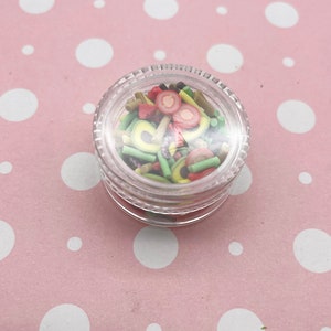 May include: A clear plastic jar filled with colorful sprinkles shaped like fruit slices and green sticks. The jar is on a pink background with white polka dots.
