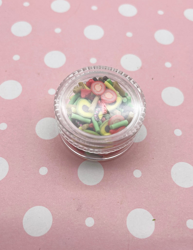 May include: A clear plastic jar filled with colorful sprinkles shaped like fruit slices and green sticks. The jar is on a pink background with white polka dots.