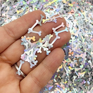 Peut inclure: Gros plan sur des paillettes argentées irisées en forme d'os. Les paillettes sont éparpillées sur une surface et une main tient une petite pile de paillettes.
