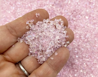 May include: A close-up of a hand holding a small amount of iridescent pink and clear glitter. The glitter is in the shape of small, irregular pieces.