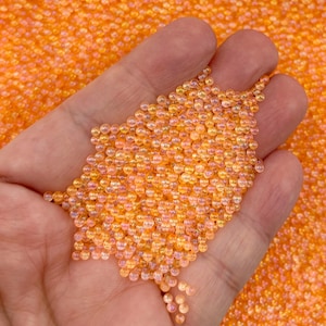 May include: Close-up of a hand holding a pile of small, translucent orange beads. The beads are spherical and have a slight iridescent sheen, reflecting light. The background is filled with similar beads, creating a textured, colorful display.