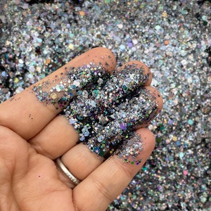 May include: A close-up of a hand holding a pile of iridescent black and silver glitter. The glitter is a mix of different sizes and shapes, and it has a holographic finish.
