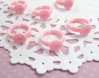 May include: A collection of six light pink plastic rings. Each ring features a circular centerpiece. The rings are arranged on a white, floral-patterned paper doily, set against a pink polka-dot background. The rings are likely children's jewelry.
