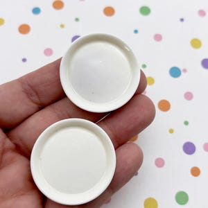 Puede incluir: Dos pequeños platos redondos de cerámica blanca se sostienen en una mano. Los platos tienen un borde ligeramente elevado y una superficie lisa y plana. El fondo es blanco con lunares de colores.