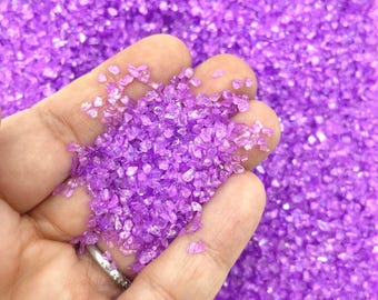 May include: A collection of small, irregular, translucent purple glass shards or crushed stone fragments. The pieces are vibrant in color, featuring a faceted texture that catches the light, creating a sparkling effect across the entire surface.