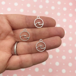 Peut inclure: Trois pendentifs de charme argentés avec un motif de vague à l'intérieur d'un cercle. Les breloques sont sur une main.