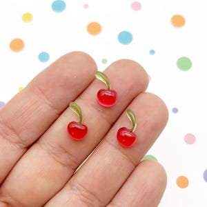 May include: Three small, translucent red cherry charms, each featuring a smooth, rounded fruit shape and a curved, light green stem. These miniature decorative pieces are crafted with a glossy finish, highlighting their vibrant color and simple design.
