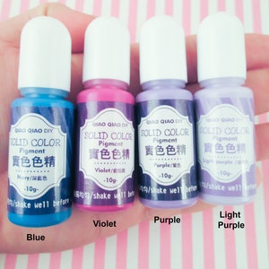 Pu&ograve; includere: Quattro piccoli flaconi di pigmento colorato solido in blu, viola, viola e viola chiaro. Ogni flacone ha un tappo bianco e un'etichetta che recita "QIAO QIAO DIY SOLID COLOR Pigment 實色色精" e il nome del colore in inglese e cinese. I flaconi sono disposti in fila da sinistra a destra, con il flacone blu a sinistra e il flacone viola chiaro a destra.