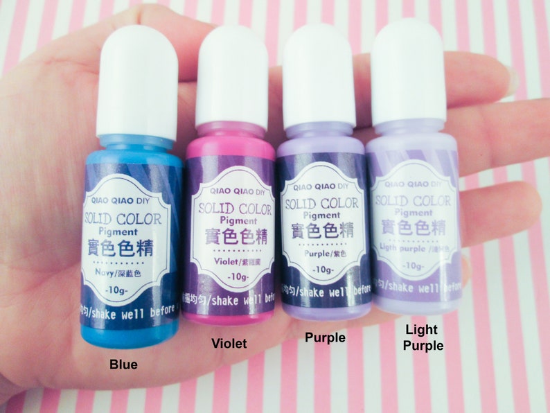 Pu&ograve; includere: Quattro piccoli flaconi di pigmento colorato solido in blu, viola, viola e viola chiaro. Ogni flacone ha un tappo bianco e un'etichetta che recita "QIAO QIAO DIY SOLID COLOR Pigment 實色色精" e il nome del colore in inglese e cinese. I flaconi sono disposti in fila da sinistra a destra, con il flacone blu a sinistra e il flacone viola chiaro a destra.