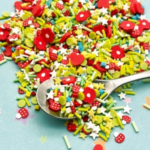 May include: A variety of colorful sprinkles in a spoon, including red hearts, yellow stars, green lines, and red flowers. The sprinkles are on a light blue background.