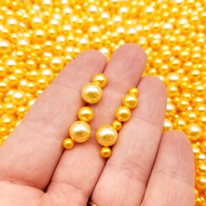 May include: Two strands of gold-colored pearl beads, each with five pearls, are held in a hand against a background of loose gold-colored pearls.