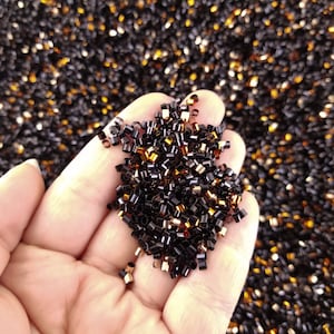 Könnte beinhalten: Nahaufnahme einer Hand, die einen Haufen kleiner, schwarzer und goldener Perlen hält. Die Perlen sind in einem zufälligen Muster angeordnet und alle etwa gleich groß.