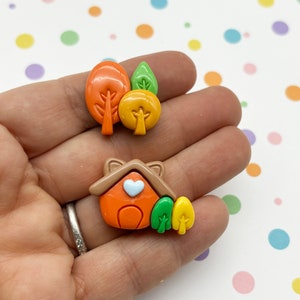 May include: A set of two clay charms featuring a small orange house with a white heart and a group of three trees in orange, yellow, and green.