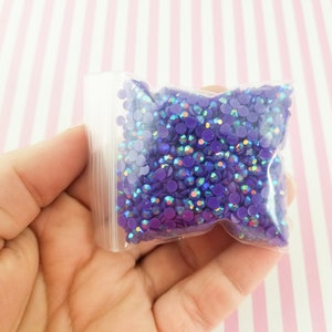 May include: A clear plastic bag filled with small, round, purple rhinestones. The rhinestones have an iridescent sheen, reflecting multiple colors. The bag is held in a hand against a pink and white striped background. These rhinestones are ideal for craft projects.