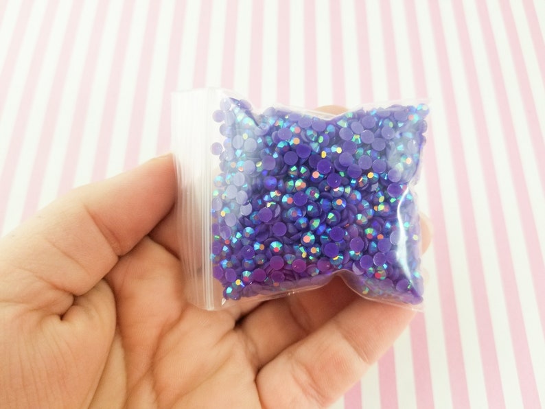 May include: A clear plastic bag filled with small, round, purple rhinestones. The rhinestones have an iridescent sheen, reflecting multiple colors. The bag is held in a hand against a pink and white striped background. These rhinestones are ideal for craft projects.