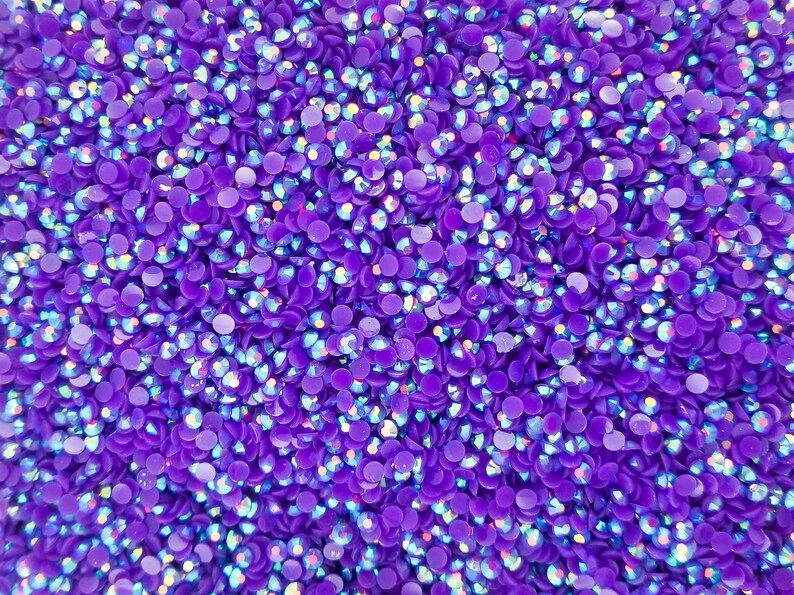 May include: Close-up of a pile of small, round, purple rhinestones. Each rhinestone has a flat back and a faceted top that reflects light, creating a shimmering effect. The rhinestones are scattered closely together.