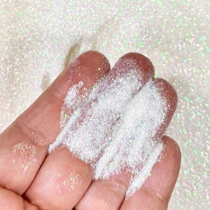 May include: Close-up of iridescent white glitter powder on fingertips. The glitter has a shimmering effect with hints of green and pink. The background is filled with the same glitter, creating a textured, sparkling effect. Ideal for crafts and decorative projects.