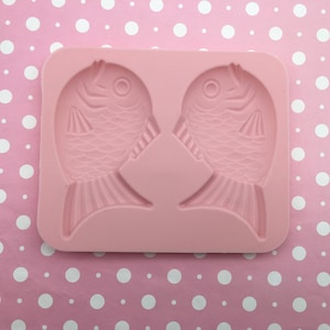 Puede incluir: Molde de silicona rosa con dos formas de pescado. El molde está diseñado para hacer dulces, chocolate u otras golosinas.