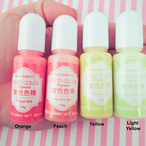 Pu&ograve; includere: Quattro piccoli flaconi di pigmento liquido in arancione, pesca, giallo e giallo chiaro. Ogni flacone ha un'etichetta bianca con il testo "QIAO QIAO DIY SOLID COLOR Pigment 實色色精" e il nome del colore in inglese e cinese. I flaconi sono disposti in fila, da sinistra a destra.