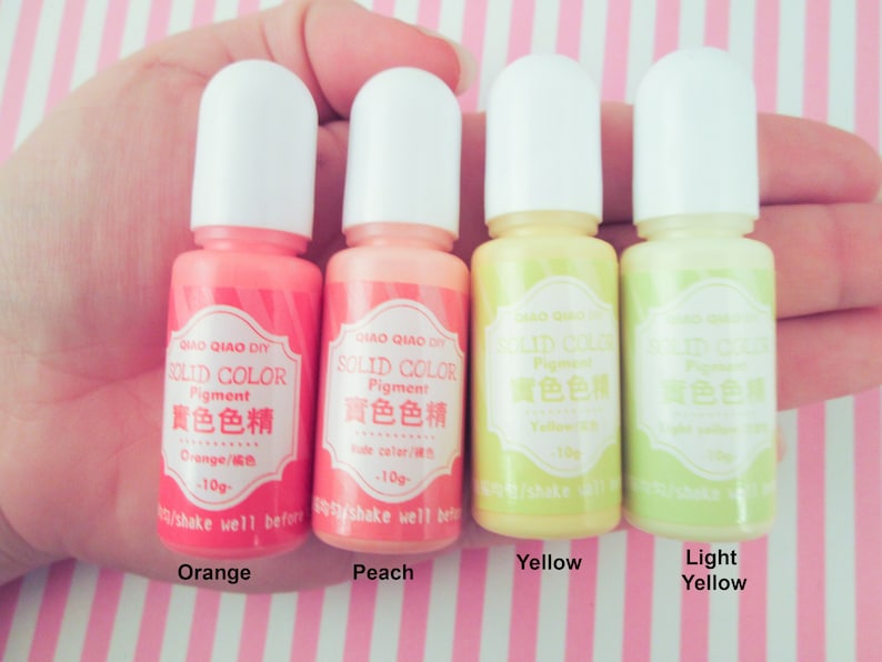 Pu&ograve; includere: Quattro piccoli flaconi di pigmento liquido in arancione, pesca, giallo e giallo chiaro. Ogni flacone ha un'etichetta bianca con il testo "QIAO QIAO DIY SOLID COLOR Pigment 實色色精" e il nome del colore in inglese e cinese. I flaconi sono disposti in fila, da sinistra a destra.