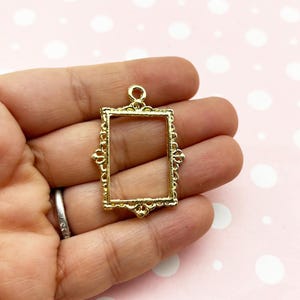 May include: Gold-toned metal picture frame pendant with an ornate design. The frame is rectangular with a small loop at the top for hanging.
