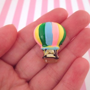 May include: A colorful hot air balloon charm with green, blue, pink, and yellow stripes. The balloon has a brown basket with a white face.