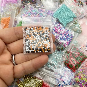 Könnte beinhalten: Eine Auswahl an kleinen, bunten Perlen in einzelnen durchsichtigen Plastiktüten. Die Perlen sind in verschiedenen Farben, darunter Schwarz, Weiß, Orange, Rot, Grün, Blau und Gold. Die Perlen sind in einem Raster angeordnet.