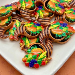 May include: A collection of colourful cornucopia-shaped decorations. Each decoration features a brown and tan striped horn overflowing with a variety of fruits and vegetables, including pumpkins, grapes, and corn. The decorations are arranged on a white rectangular plate.