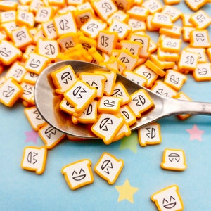 May include: A spoon filled with small yellow and white plastic charms shaped like slices of bread with a smiling face. The charms are scattered on a blue surface with yellow stars.