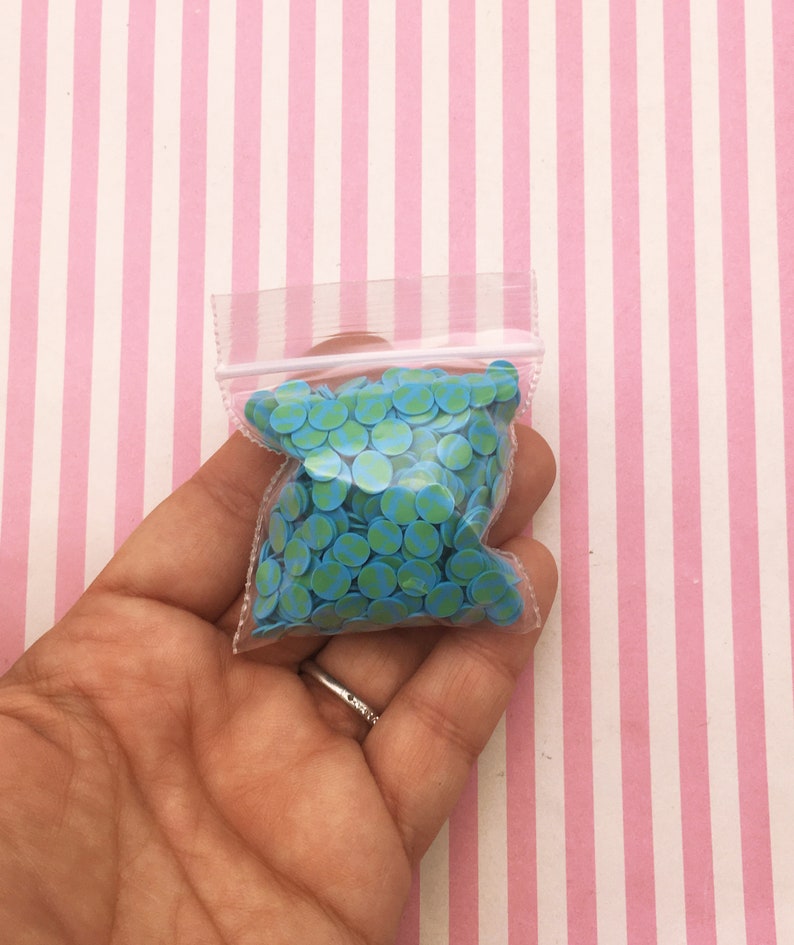 May include: A small clear plastic bag filled with blue and green round sprinkles. The sprinkles have a green and white pattern.
